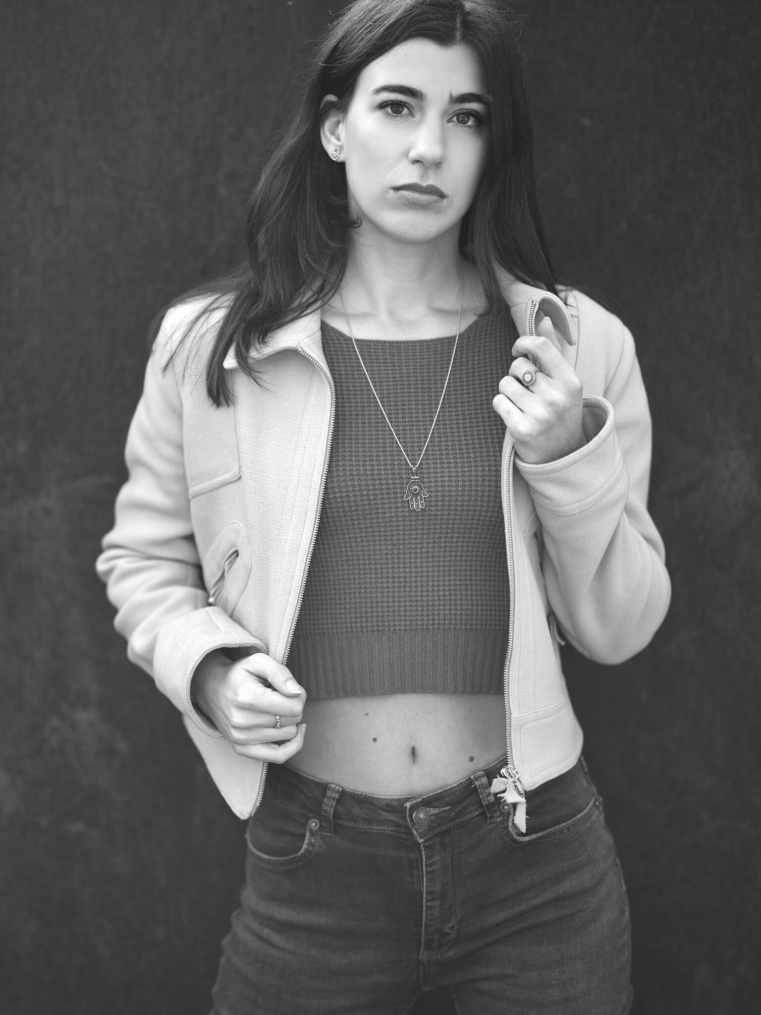 Auto-generated description: A person stands against a dark background wearing a jacket over a crop top with a pendant necklace.