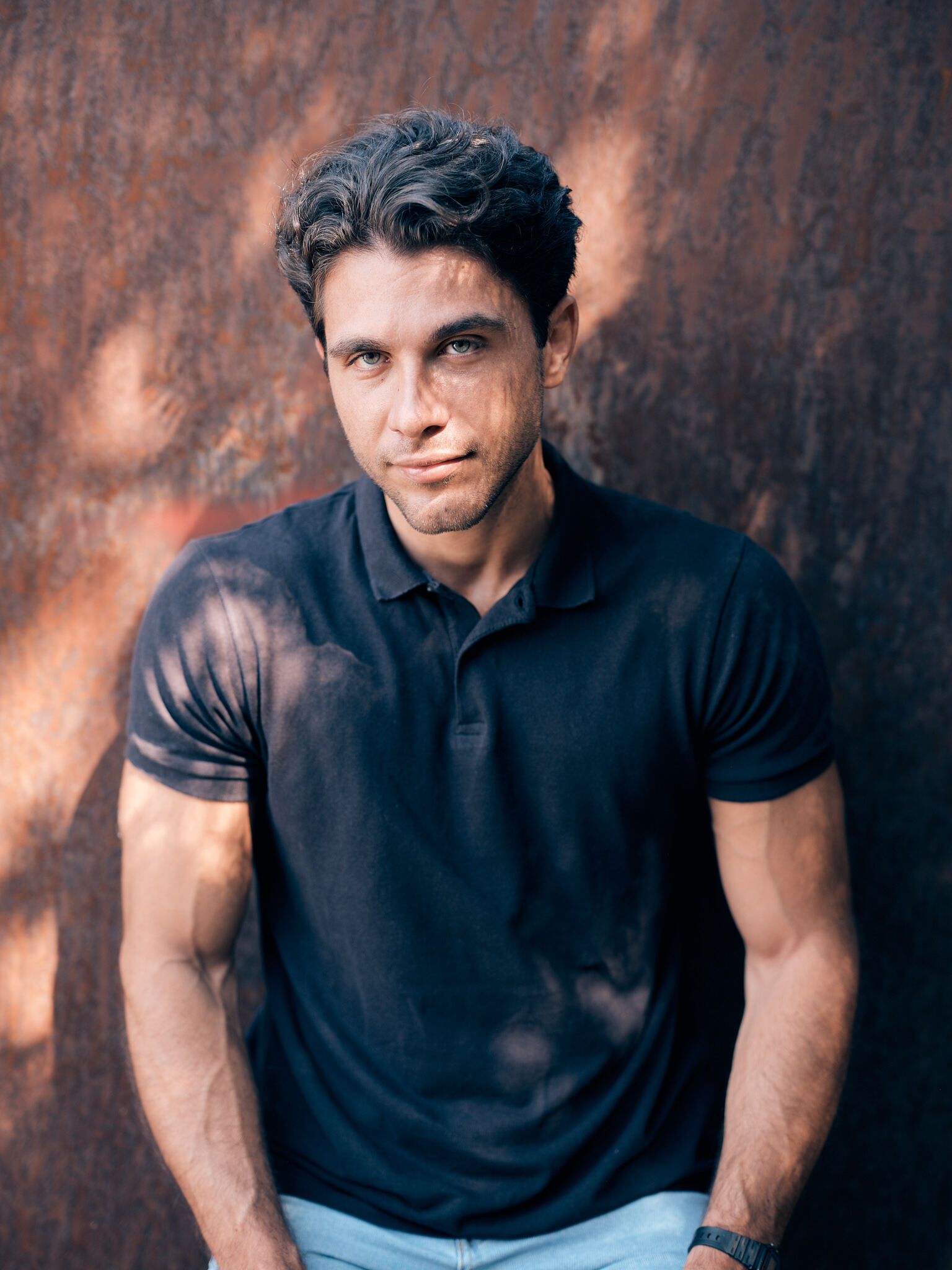 A young man with wavy hair and a confident expression is wearing a black polo shirt and standing against a textured, reddish-brown wall with light and shadow patterns.
