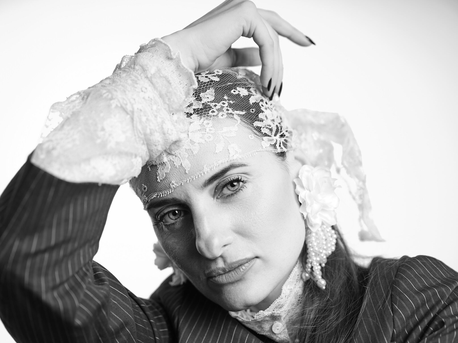 A person with a lace head covering and elaborate earrings poses with one hand resting on their head, gazing directly at the camera.
