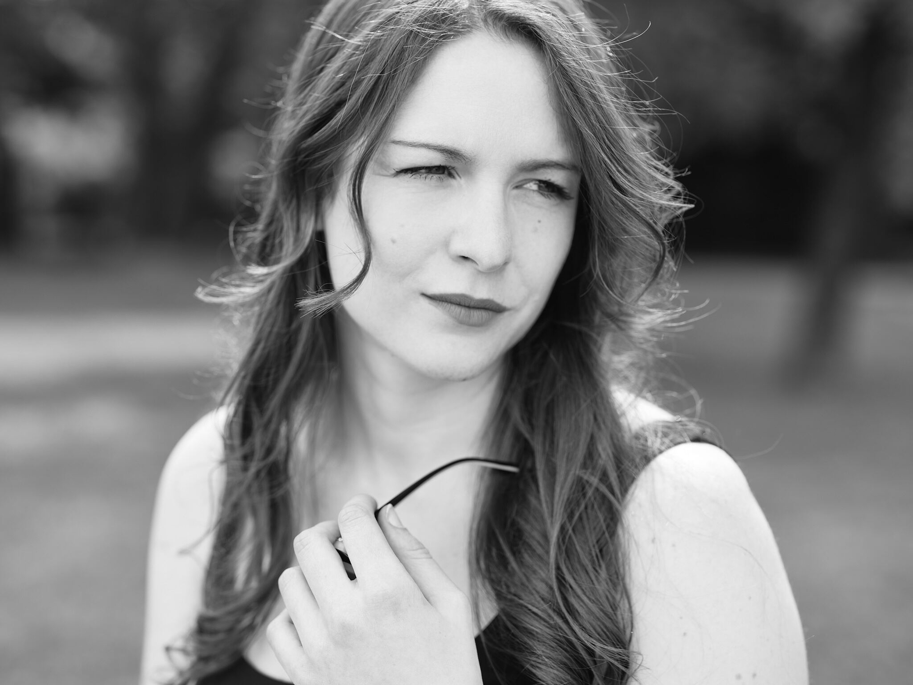 A woman with long hair is holding a pair of glasses and looking thoughtfully to the side.