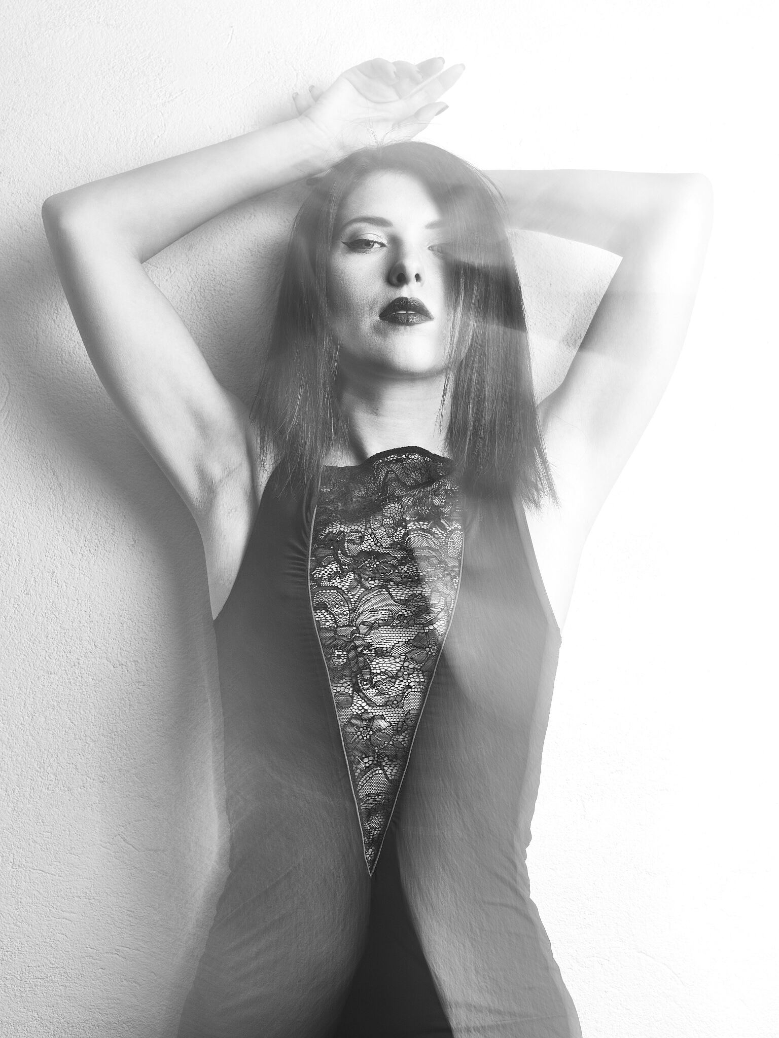 A woman in a sleeveless lace dress poses with her arms raised above her head against a plain wall, captured with a motion blur effect.